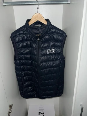 Svart dunväst från EA7 Emporio Armani - Snygg svart dunväst från EA7 Emporio Armani med quiltad design och hög krage. Västen har dragkedja framtill och en stilren logga på bröstet. Västen är i storlek XXL men passar även för XL. Perfekt för lager på lager och ger en sportig vibe till din outfit.