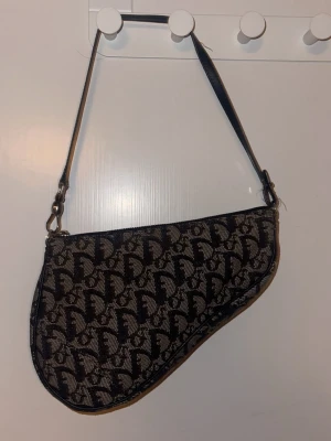 Dior Saddle Bag i svart och beige - Klassisk Dior Saddle Bag i svart och beige canvas med det klassiska Dior-monogrammet över hela väskan. Väskan har en unik sadelform. Perfekt statement piece för dig som vill sticka ut. Skriv om frågor eller om fler bilder. ❤️Pris kan diskuteras!