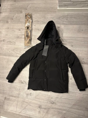 Svart dunjacka från Canada Goose - Snygg svart dunjacka från Canada Goose med avtagbar huva och tillhörande pälskant. Jackan har stora fickor framtill, dragkedja och tryckknappar. Perfekt för kalla vinterdagar och har en stilren, modern look.