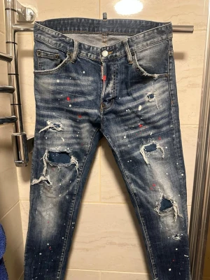 Dsquared2 jeans - Säljer ett par mörkblå jeans med skinny fit, slitna detaljer och hål på benen. Jeansen har färgstänk i vitt och rött.