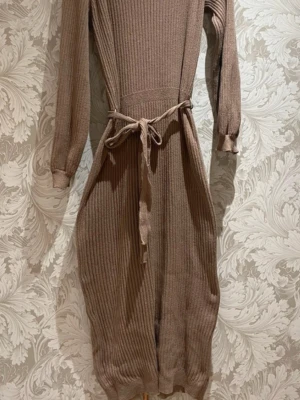 Beige stickad klänning från Bubbleroom - Mysig beige stickad klänning från Bubbleroom med lång ärm, ribbad struktur och knytskärp i midjan. Perfekt för dig som gillar en avslappnad men ändå snygg stil. Klänningen har en rak passform och är knälång.