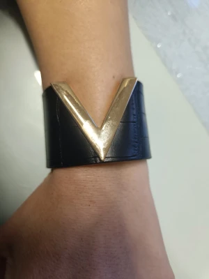 Svart läderarmband med gulddetalj - Stilrent svart armband i läder med en stor V-formad guldfärgad metall-detalj framtill. Bred design som ger ett statement på handleden. Perfekt för dig som gillar accessoarer med attityd och modern känsla. Mått 21x4 cm   nytt. Oanvänd 