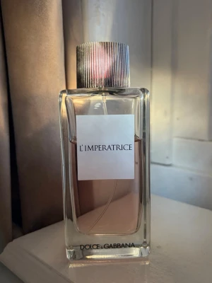 Dolce & Gabbana L'Imperatrice parfym - Dolce & Gabbana L'Imperatrice är en elegant parfym i en stilren, rektangulär glasflaska med silverfärgad räfflad kork. Doften har en ljusrosa ton och flaskan har en minimalistisk vit etikett framtill.