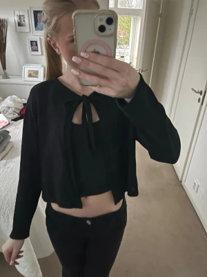 Svart croppad topp med knyt - Svart croppad topp med lång ärm och snygg knytning framtill vid halsen. Toppen har en öppen detalj vid bröstet och är tillverkad i ett mjukt material som känns bekvämt. Perfekt för dig som gillar trendiga och stilrena plagg.