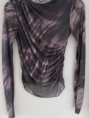 Långärmad mesh-topp med drapering - Långärmad topp i stretchig mesh med gråsvart mönster. Transparent material med rynk/drapering framtill som ger fin passform. Rund hals med knappstängning bak. Lätt och följsam i tyget.  Passar perfekt till jeans, kjol eller över linne/bh.  Fint skick.  Storlek: S