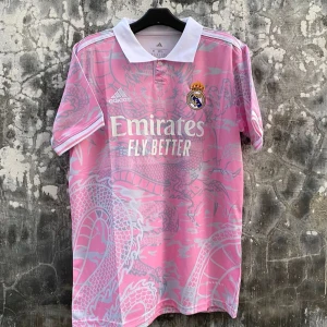 Rosa Real Madrid träningströja Adidas - Säljer en rosa Real Madrid träningströja från Adidas med vit krage och drakmönster i ljusgrått. Tröjan har korta ärmar, klubbmärke på bröstet och tryck med 'Emirates Fly Better' framtill. Perfekt för fotbollsträning eller som supporterplagg.