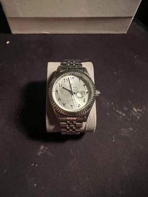 Silverfärgad klocka med arabiska siffror - Stilren klocka från Geneva i silverfärg med metallarmband och räfflad boett. Urtavlan är vit med arabiska siffror och datumvisning. Klassisk design som passar till många stilar. Fungerar felfritt