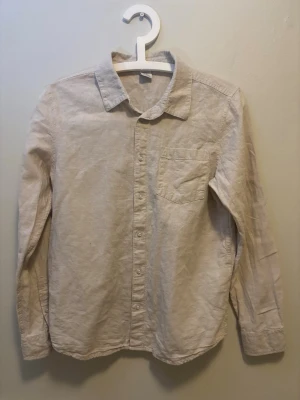 Beige skjorta från Lindex Kids - Stilren beige skjorta från Lindex Kids i storlek 158. Skjortan har klassisk krage, bröstficka och knäppning framtill. Tillverkad i ett mjukt material som känns skönt mot huden. Perfekt för dig som gillar en enkel och clean look.