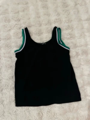 Svart linnetopp med gröna detaljer - Snygg svart linnetopp med gröna och vita ribbade kanter vid axelbanden. Perfekt för en sportig och avslappnad stil. Materialet känns mjukt och stretchigt, vilket gör toppen bekväm att bära hela dagen accommodating olika looks.