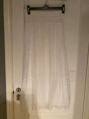 Vit långkjol från Gina Tricot - Luftig och flowy vit långkjol från Gina Tricot med smock i midjan. Kjolen har flera volanger och är tillverkad i ett lätt material som passar perfekt till varmare dagar.