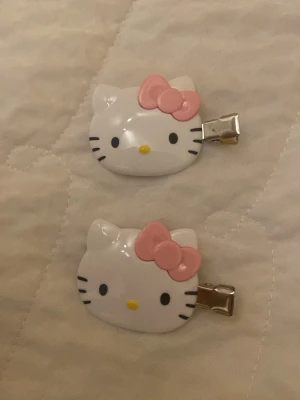 Hello Kitty hårspännen med rosa rosett - Två söta hårspännen med Hello Kitty-motiv i vitt och svart, dekorerade med en rosa rosett. Spännena är gjorda i plast med metallklämma på baksidan. Perfekta för att piffa upp frisyren med en gullig detalj.