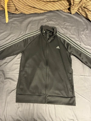 Svart Adidas träningsjacka - Snygg svart träningsjacka från Adidas med dragkedja framtill och klassiska tre ränder i grått längs ärmarna. Jackan har hög krage, två sidofickor och Adidas-logga på bröstet. Perfekt för gymmet eller vardagslooken.