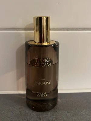 Zara Tonka Cream Eau de Parfum - Zara Tonka Cream Eau de Parfum i en elegant brun glasflaska med guldfärgat lock. Flaskan har en rundad form och stilren design med text i guld. Perfekt för dig som gillar lyxiga och moderna dofter.