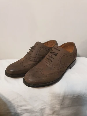 Bruna loafers i läder med broguemönster - Säljer ett par bruna loafers i äkta läder med klassiskt broguemönster och rundad tå. Skorna har låg klack, dekorativa perforeringar och snörning framtill. Perfekta för dig som gillar stilrena och tidlösa skor med en twist. Storleken är 7 1/2, vilket är 40-41