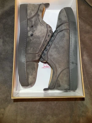 Gråa Louboutin sneakers i mocka - Snygga gråa sneakers från Christian Louboutin i mjuk mocka med klassisk röd sula. Lågt skaft och diskret design som ändå sticker ut med den ikoniska röda undersidan. Perfekta för dig som gillar exklusiva detaljer.