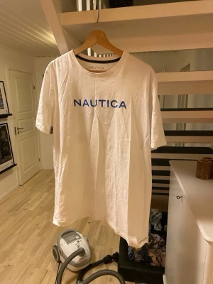 Vit Nautica t-shirt med tryck - Vit t-shirt från Nautica med blå logotyp framtill och stort grafiskt tryck på ryggen. Klassisk rund hals och korta ärmar. Tillverkad i mjuk bomull för en skön känsla. Perfekt för dig som gillar stilrena och sportiga plagg.