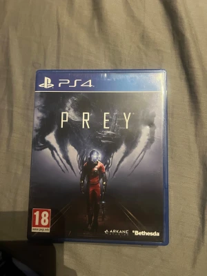 Prey till PS4 - Prey till PlayStation 4, ett sci-fi actionspel från Arkane Studios och Bethesda. Spelet levereras i blå plastfodral med omslag och skiva. Omslaget har en astronaut i röd dräkt och mörka, mystiska former i bakgrunden.