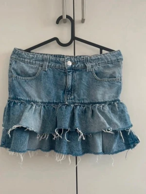 Blå jeanskjol med volanger - Säljer en blå jeanskjol med volangdetaljer och råa kanter. Kjolen har klassisk femficksdesign, bälteshällor och knäppning fram. Perfekt för dig som gillar en avslappnad och trendig stil.