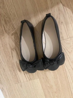 Svarta ballerinaskor med rosett - Stilrena svarta ballerinaskor med stor dekorativ rosett framtill. Skorna har rundad tå och är tillverkade i ett slätt material som ger en klassisk look. Perfekta för dig som gillar enkel men söt stil.