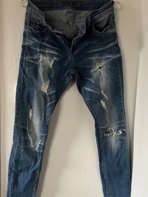 Blå slitna jeans med distressed detaljer - Säljer ett par blå jeans från H&M med skinny fit och tydliga distressed detaljer och slitningar framtill. Jeansen har klassisk femficksmodell, normal midja och är tillverkade i denim med stretch. Perfekta för en avslappnad och trendig look.