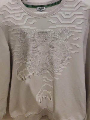 Vit sweatshirt från Kenzo med tiger - Säljer en vit sweatshirt från Kenzo med det ikoniska tigermotivet broderat framtill och coola 3D-detaljer både fram och bak. Tröjan har rund halsringning, långa ärmar och är gjord i mjukt bomullsmaterial. Perfekt för dig som vill sticka ut med stilren design.