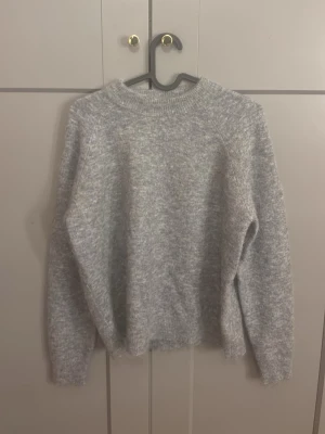Grå fluffig stickad tröja - Mysig grå stickad tröja med rund hals och ribbade muddar. Tröjan har en fluffig och mjuk känsla, perfekt för kyliga dagar. Passar dig som gillar en enkel men stilren look. Materialet ser ut att vara en ull- eller mohairblandning.