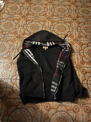 Svart hoodie med dragkedja Burberry - Svart hoodie från Burberry med dragkedja och klassiskt rutmönster på insidan av huvan. Hoodien har ribbade muddar, fickor på sidorna och är tillverkad i mjukt material. Perfekt för dig som vill ha en stilren och exklusiv look.