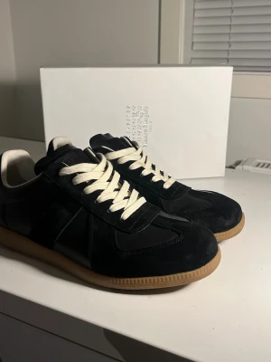 Svarta Maison Margiela sneakers - Maison Margiela sneakers i svart läder och mocka med vita skosnören och beige gummisula. Klassisk låg modell med diskret logotyp på plösen och vit häl. Snygg och stilren design som passar till många outfits. Skickar samma dag!
