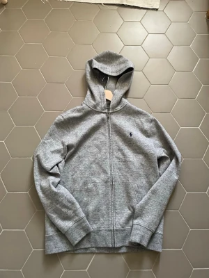Grå hoodie från Polo Ralph Lauren - Klassisk grå hoodie med dragkedja från Polo Ralph Lauren. Tröjan har huva, två fickor framtill och den ikoniska mörkblå loggan broderad på bröstet. Tillverkad i mjukt sweatshirtmaterial som är perfekt för chill dagar.