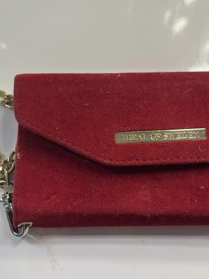 Rött sammetsskal Ideal of Sweden IPhone 6 - Snyggt rött mobilskal i sammet från Ideal of Sweden med guldfärgad kedja och logga. Plånboksmodell med magnetstängning och plats för kort. Perfekt accessoar för dig som vill ha både stil och funktion.