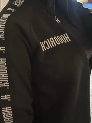 Svart track jacket från Hoodrich - Svart track jacket från Hoodrich med vit logga tryckt på bröstet och längs ärmarna. Jackan har hög krage och dragkedja framtill. Sportig och stilren design, perfekt för träning eller streetwear.