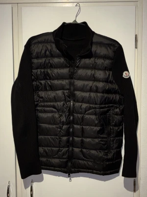 Svart dunväst från Moncler - Snygg svart dunväst från Moncler med stickade ärmar och hög krage. Västen har quiltad front, dragkedja och två fickor framtill. Moncler-logga på vänster ärm ger en exklusiv touch. Perfekt för lager-på-lager och streetstyle.
