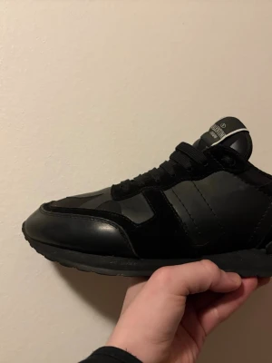 Svarta sneakers från Valentino - Snygga helsvarta sneakers från Valentino med coola nitar på hälen. Skorna har en mix av mocka och läderdetaljer, samt svarta skosnören. Perfekta för dig som gillar stilrena och exklusiva sneakers med en edgy touch.