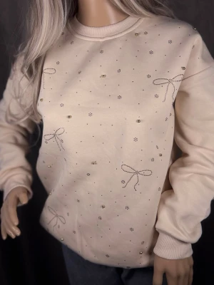 Beige sweatshirt med pärlor och rosetter - Supersöt beige sweatshirt med små pärlor och broderade rosetter över hela framsidan. Tröjan har rund halsringning, ribbade muddar och en avslappnad passform. Perfekt för dig som gillar detaljer och en mjuk, bekväm stil.