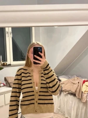 Beige och svart randig kofta - Mysig beige kofta med svarta horisontella ränder och v-ringning. Koftan har knappar framtill, ribbade muddar och är något croppad i modellen. Perfekt att slänga över ett linne eller t-shirt för en avslappnad look.