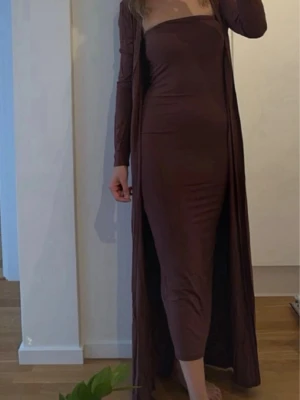Brun axelbandslös långklänning med cardiga - Snygg brun axelbandslös långklänning med figurnära passform. Klänningen har en enkel och stilren design och är tillverkad i ett stretchigt material som sitter bekvämt. Perfekt för dig som gillar minimalistisk stil.