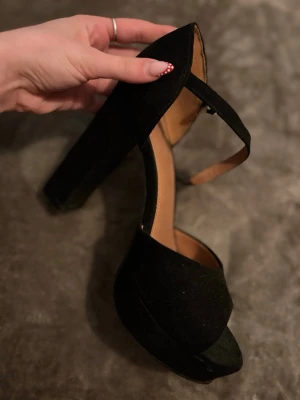 Svarta chunky pumps med ankelrem - Säljer ett par svarta pumps med bred klack och öppen tå. Skorna har en snygg ankelrem med spänne och är klädda i mockaimitation. Perfekta för dig som gillar en stilren men trendig look. Lite slitna vid spännena!