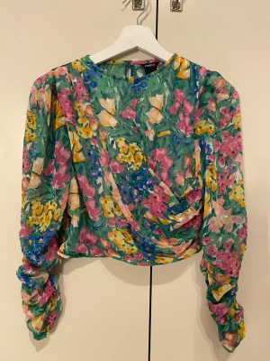 Blommig långärmad blus från Lindex - Färgglad blus från Lindex med grönt, rosa, gult och blått blommönster. Modellen har långa, rynkade ärmar och en draperad front. Tillverkad i ett tunt, transparent material som ger en luftig känsla. Perfekt för dig som gillar statement-plagg och unika prints.