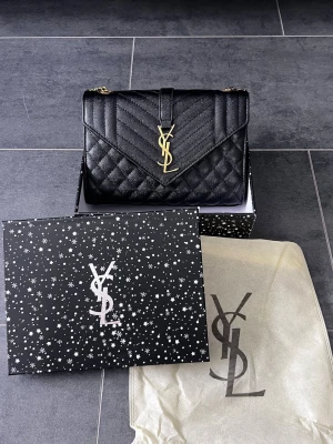 Svart handväska från YSL - Snygg svart handväska från Yves Saint Laurent i quiltat läder med guldigt YSL-emblem framtill. Väskan har kedjerem i guld och klassisk kuvertform. Kommer med original ask och dustbag med YSL-logga. Perfekt för dig som vill ha en lyxig accessoar.