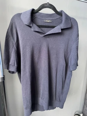 Mörkblå stickad pikétröja Massimo Dutti - Snygg mörkblå stickad pikétröja från Massimo Dutti med korta ärmar och klassisk krage. Tröjan har en avslappnad passform och är tillverkad i ett mjukt, tunt material som känns skönt mot huden. Perfekt för en stilren och enkel look.