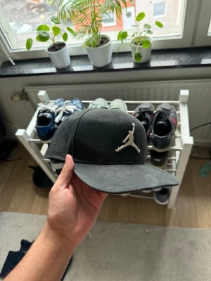 Svart Jordan keps med broderad logga - Säljer en svart keps från Jordan med den klassiska Jumpman-loggan broderad i vitt framtill. Kepsen har en rak skärm och är tillverkad i ett mjukt tygmaterial. Perfekt för dig som gillar streetwear och sportig stil.