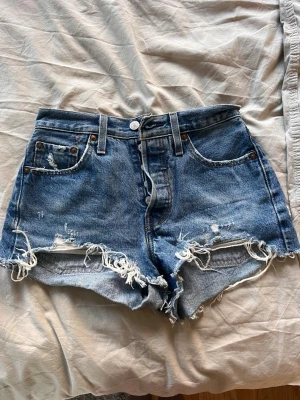 Levi's blåa jeansshorts med fransar - Snygga blå jeansshorts från Levi's med hög midja och råa, fransiga kanter. Klassisk femficksmodell med slitna detaljer och knappgylf. Perfekta för sommaren och ger en cool, avslappnad vibe.