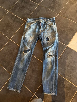 Dsquared2 slitna blå jeans - Säljer ett par blå jeans från Dsquared2 med coola slitningar och hål på benen. Jeansen har en smal passform, klassisk femficksdesign och detaljer som färgstänk och tvättad look. Perfekta för dig som gillar en edgy streetstil. Dom är helt äkta. Skriv så kan vi förhandla om pris