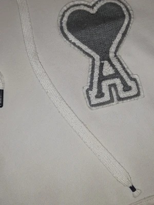 Beige hoodie från Ami Paris med hjärtdetalj - Säljer en beige hoodie från Ami Paris med den ikoniska hjärta-A-loggan i grått och vitt på bröstet. Hoodien har dragsko i huvan och är gjord i mjukt bomullsmaterial. Perfekt för dig som gillar stilrena och trendiga plagg.