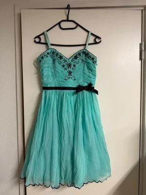 Turkos kortklänning med broderi - Söt turkos kortklänning med svarta axelband, svart broderi med blommor upptill och en svart rosett i midjan. Klänningen har v-ringad urringning och ett lätt, svepande tyg som ger volym. Perfekt för dig som vill sticka ut med en färgglad look. En liten fläck nedre delen av klänningen. Köpt på ASOS.