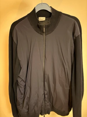 Svart ziptröja från John Henric - Stilren svart ziptröja från John Henric med stickade ärmar och rygg samt slät framsida i ett annat material. Hög krage och hel dragkedja framtill. Perfekt för lager-på-lager och en clean look.