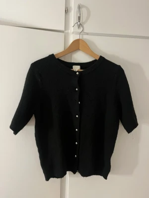 Svart kortärmad kofta med pärlknappar - Snygg svart kofta med korta ärmar och rund halsringning. Koftan har pärlknappar framtill som ger en elegant touch. Perfekt att styla över en topp eller klänning för en trendig look. Bara använda några gånger men är väldigt mjuk och skön.