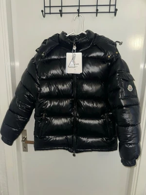 Svart dunjacka från Moncler - Säljer min Moncler dunjacka i svart. Jackan är i mycket fint skick och kommer självklart med originala etiketter, serienummer och autentiserings-QR-kod, vilket gör det enkelt för köparen att verifiera äktheten via code.moncler.com.  ✔ Äkta – QR-kod & certilogo-etikett ✔ Fint skick – inga fläckar eller skador ✔ Varm, lätt och perfekt för både vardag och vinter ✔ Klassisk Moncler-design med ikoniska serietidnings-fodret  Storlek: S Färg: Svart Köpt: 2026