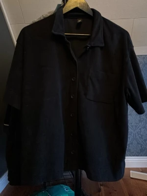 Svart kortärmad skjorta med knappar - Säljer en svart pike från Weekday med korta ärmar och klassisk krage. Skjortan har knappar framtill och en bröstficka. Perfekt för dig som gillar en enkel och stilren look.