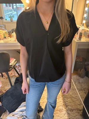 Svart kortärmad blus med krage - En stilren svart blus med korta ärmar och v-ringad krage. Blusen har en lös passform och är tillverkad i ett lätt material som ger en snygg och avslappnad look. Perfekt att matcha med jeans eller kjol för en trendig outfit.